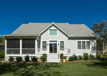 A house sits far off the curb with white siding and a gray roof. There are trees growing on the right side. There's a porch on the left side.