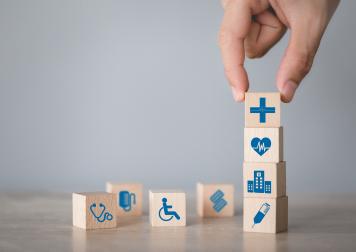 Blocks with health insurance symbols on them. A hand comes from the top to place block on top of stack of blocks.